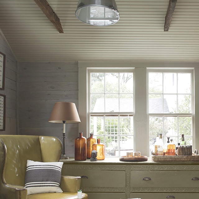 A comfortable sitting area with soft, neutral tones, shiplap walls, a light gray-painted beadboard ceiling and trim, a light-sage green dresser under two windows, and a yellow leather chair. A comfortable sitting area with soft, neutral tones, shiplap walls, a light gray-painted beadboard ceiling and trim, a light-sage green dresser under two windows, and a yellow leather chair.