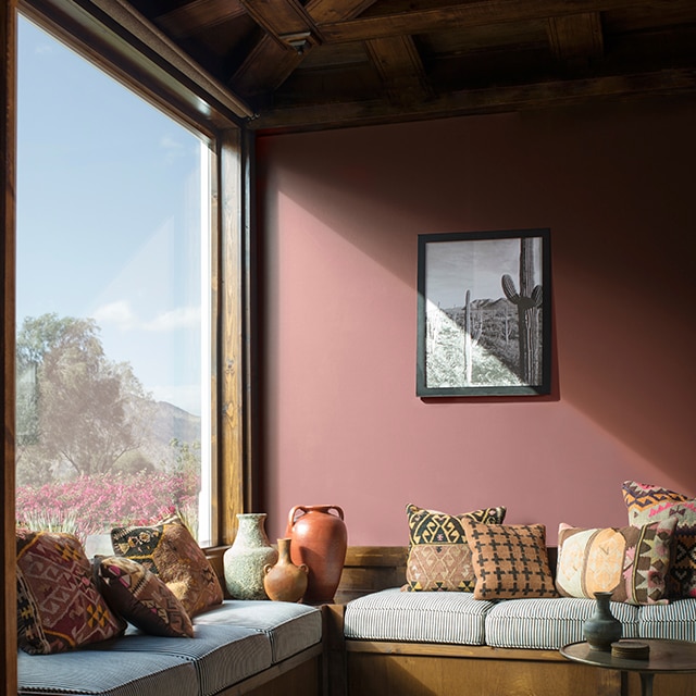 Sun streaming into a muted, brick red-painted living room with a wraparound window seat, southwestern style décor, and a wood beamed ceiling. Sun streaming into a muted, brick red-painted living room with a wraparound window seat, southwestern style décor, and a wood beamed ceiling.