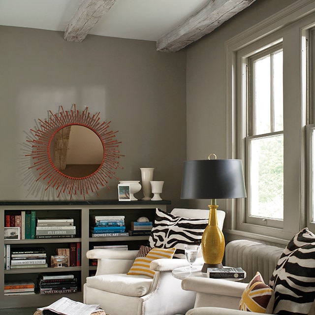 A cozy living room with deep taupe-painted walls, trim, and low built-in bookcase, two white chairs with zebra print pillows, and a white ceiling with wood beams. A cozy living room with deep taupe-painted walls, trim, and low built-in bookcase, two white chairs with zebra print pillows, and a white ceiling with wood beams.