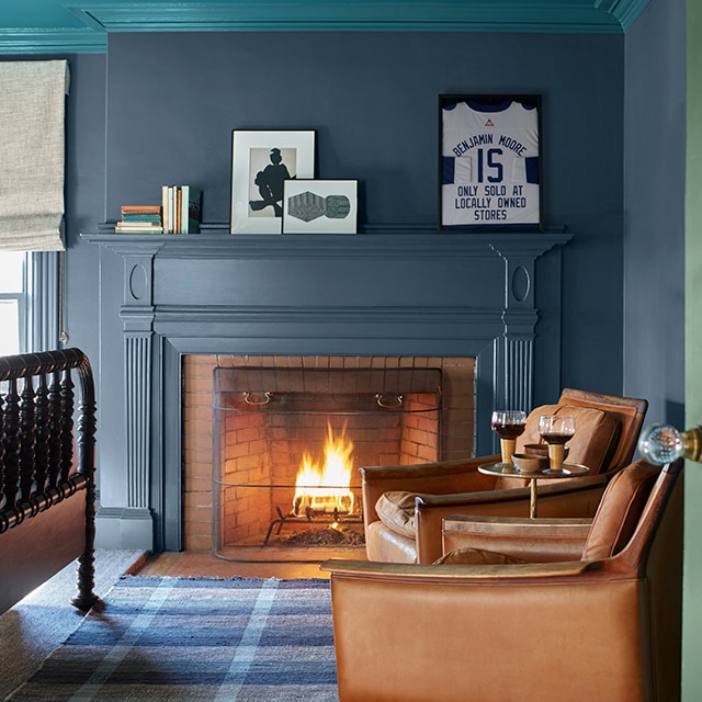 A cozy corner of a room with dark teal-blue painted walls and working fireplace mantel, a teal ceiling and crown molding, two brown leather chairs, a blue plaid rug, and the edge of a green door. A cozy corner of a room with dark teal-blue painted walls and working fireplace mantel, a teal ceiling and crown molding, two brown leather chairs, a blue plaid rug, and the edge of a green door.