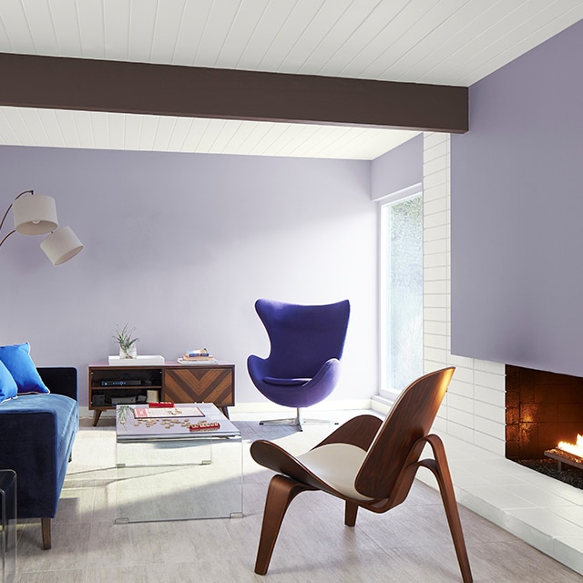 A bright, modern living room with light violet painted walls and accent wall over the fireplace, white shiplap walls and ceiling, a dark brown ceiling beam, navy sofa and purple egg chair. A bright, modern living room with light violet painted walls and accent wall over the fireplace, white shiplap walls and ceiling, a dark brown ceiling beam, navy sofa and purple egg chair.