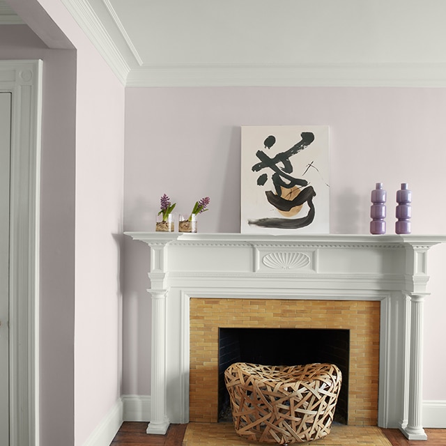 Pretty light violet-painted corner walls, white crown molding, trim and fireplace mantel with modern artwork and purple vases, and a white hallway door on the left. Pretty light violet-painted corner walls, white crown molding, trim and fireplace mantel with modern artwork and purple vases, and a white hallway door on the left.