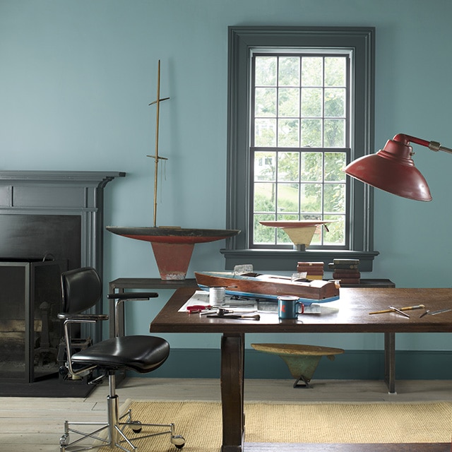 A spacious home office with blue-gray painted walls, a dark gray mantel and trim, a large wood desk, leather swivel chair, red floor lamp, and wooden toy boats. A spacious home office with blue-gray painted walls, a dark gray mantel and trim, a large wood desk, leather swivel chair, red floor lamp, and wooden toy boats.