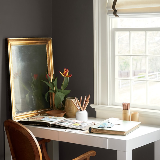 A pretty corner home office space with chocolate brown painted walls and a large, white-trimmed window in front of a white desk with scrapbooks and pencils, and a wood chair. A pretty corner home office space with chocolate brown painted walls and a large, white-trimmed window in front of a white desk with scrapbooks and pencils, and a wood chair.