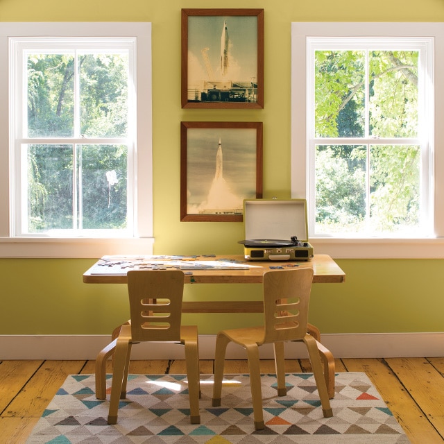 A children’s wooden desk, two chairs, and a multicolor rug in front of a light green painted wall with white trim and rocket pictures flanked by two windows. A children’s wooden desk, two chairs, and a multicolor rug in front of a light green painted wall with white trim and rocket pictures flanked by two windows.