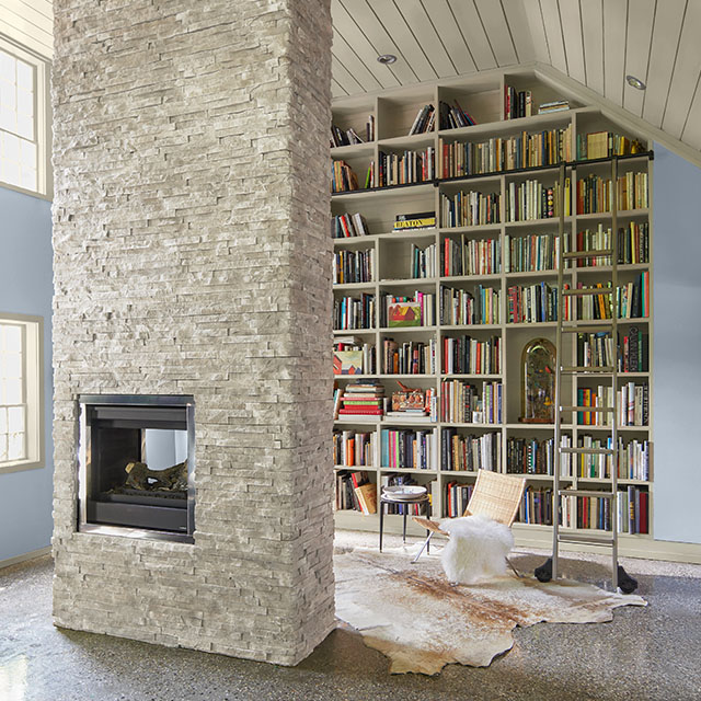 A home library with a statement floor-to-ceiling white-painted bookcase, blue walls, white shiplap vaulted ceiling and trim, and white stone center fireplace. A home library with a statement floor-to-ceiling white-painted bookcase, blue walls, white shiplap vaulted ceiling and trim, and white stone center fireplace.