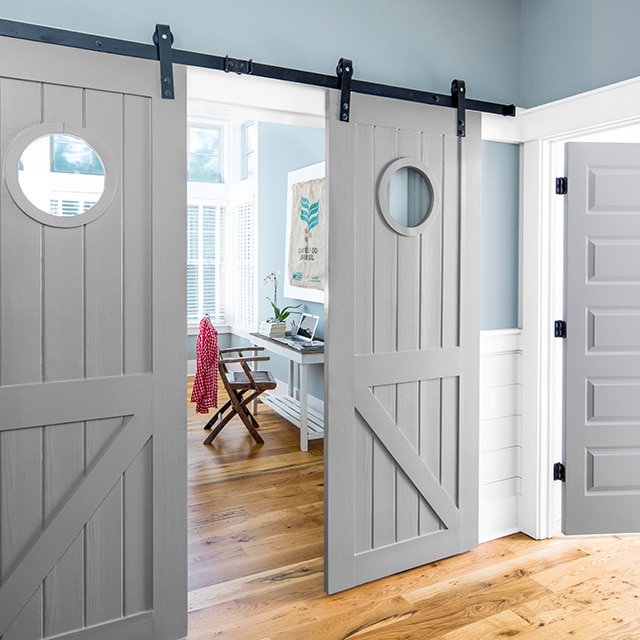 A pretty, blue painted hallway looking into a blue painted office space with a small white desk, a brown chair, white trim, and open gray painted sliding barn doors. A pretty, blue painted hallway looking into a blue painted office space with a small white desk, a brown chair, white trim, and open gray painted sliding barn doors.