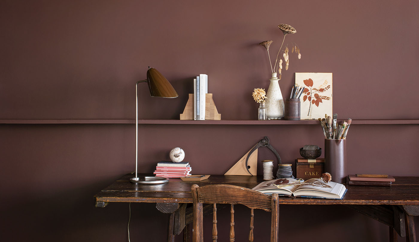 A rich, dark brown painted wall with a long shelf holding knickknacks, behind a rustic wood desk and chair. A rich, dark brown painted wall with a long shelf holding knickknacks, behind a rustic wood desk and chair.