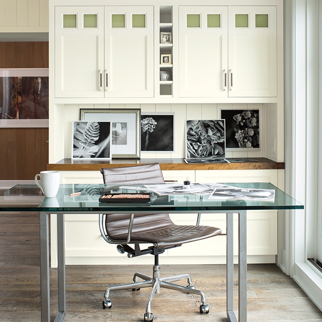 A bright, airy, white painted home office with shiplap walls and white built-in cabinets behind a glass-top desk and brown swivel chair. A bright, airy, white painted home office with shiplap walls and white built-in cabinets behind a glass-top desk and brown swivel chair.