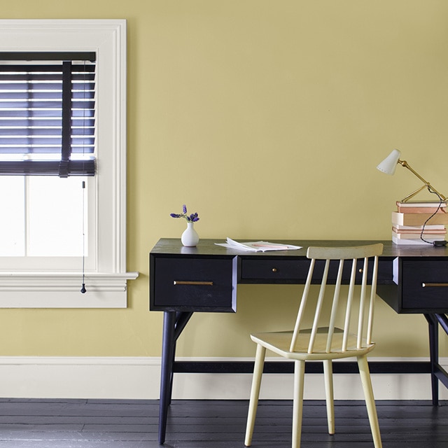 A home office area with a black desk and yellow chair against a soft yellow painted wall with white trim, and a window with black open blinds. A home office area with a black desk and yellow chair against a soft yellow painted wall with white trim, and a window with black open blinds.