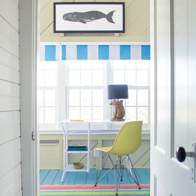 A white painted hallway wall and door open into a coastal vibe home office with a pale-yellow painted shiplap wall and ceiling, a framed whale painting hanging over windows, a white desk and yellow chair, and a blue painted floor. A white painted hallway wall and door open into a coastal vibe home office with a pale-yellow painted shiplap wall and ceiling, a framed whale painting hanging over windows, a white desk and yellow chair, and a blue painted floor.
