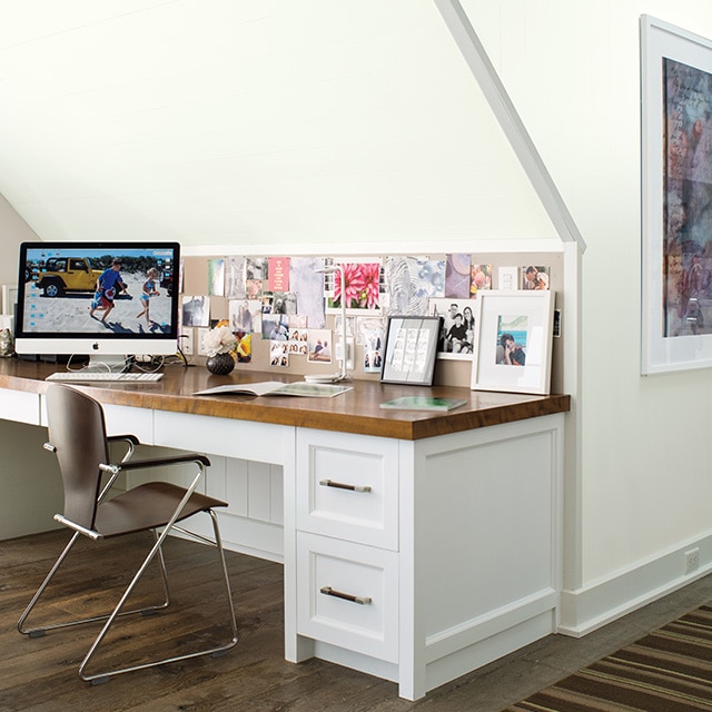 A white painted built-in desk with a wood top, a computer, and pictures beneath a white sloped wall, and a brown metal chair. A white painted built-in desk with a wood top, a computer, and pictures beneath a white sloped wall, and a brown metal chair.