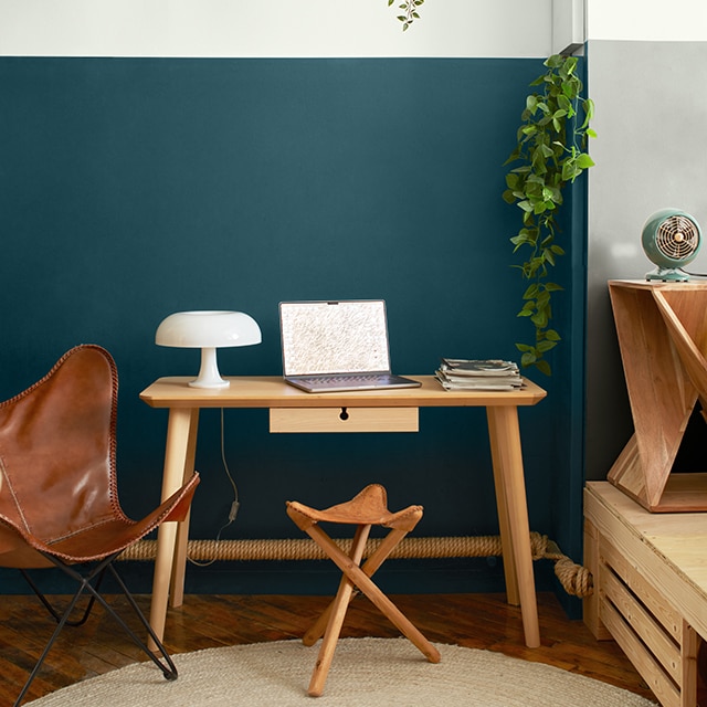 A wood desk, leather butterfly chair, and stool in front of a two-tone white and teal blue painted wall with hanging plants on an upper shelf, and an adjacent white and gray two-tone wall. A wood desk, leather butterfly chair, and stool in front of a two-tone white and teal blue painted wall with hanging plants on an upper shelf, and an adjacent white and gray two-tone wall.