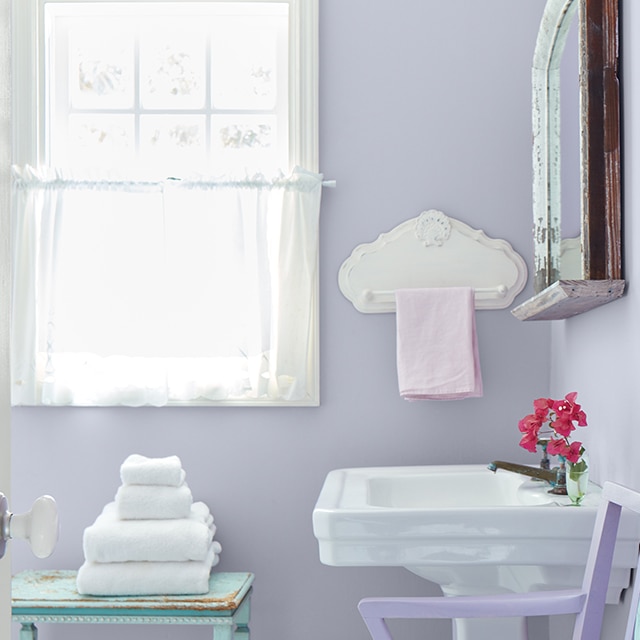 A sunny small bathroom with pastel purple walls, a white sink, and a white-trimmed window with gauzy white curtains. A sunny small bathroom with pastel purple walls, a white sink, and a white-trimmed window with gauzy white curtains.