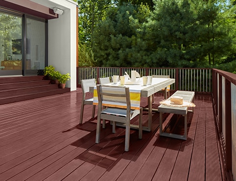 A burgundy red stained deck with outdoor dining furniture and stained steps leading to a home’s large sliding glass doors. A burgundy red stained deck with outdoor dining furniture and stained steps leading to a home’s large sliding glass doors.