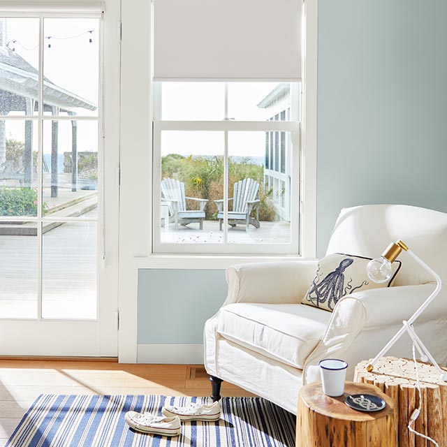 A sunny, light blue painted living room with a cozy white armchair, and an ocean view through a glass door and window.