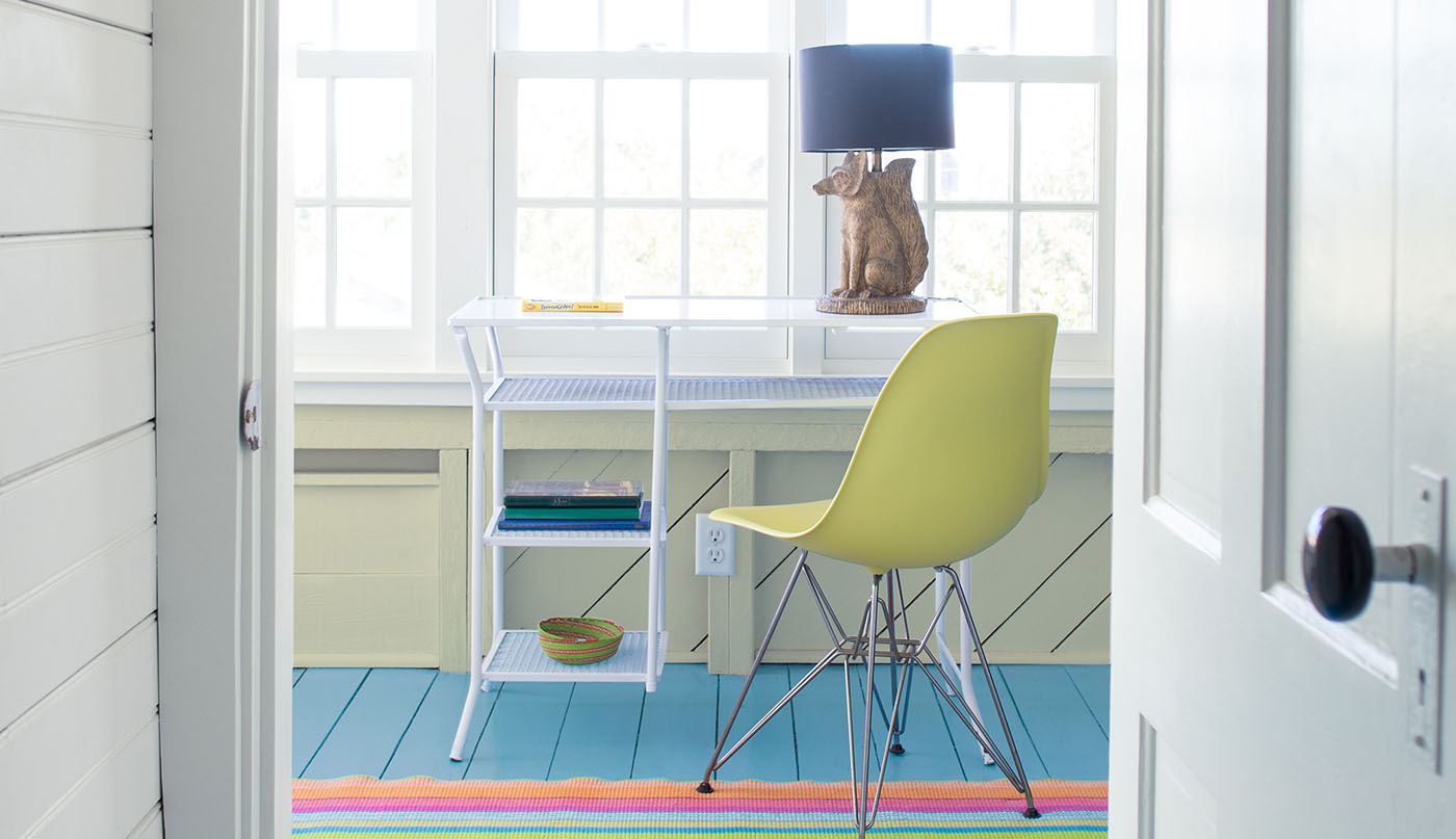 A white painted hallway wall and door open into a coastal vibe home office with a pale-yellow painted shiplap wall and ceiling, a framed whale painting hanging over windows, a white desk and yellow chair, and a blue painted floor. 
