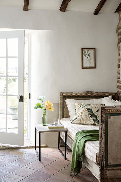 A white room with a wood beam ceiling, a daybed, and a door propped open leading outside.