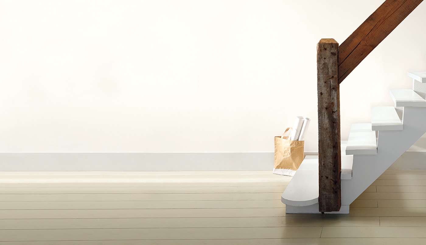 A rustic white staircase with wooden handrail in an off-white room; a tote filled with rolled posters sits on the floor.