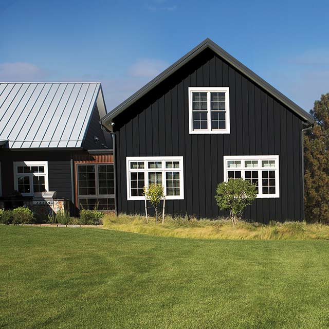 A black farmhouse in a field features white windows, surrounded by mountains and red flowers on a sunny day. A black farmhouse in a field features white windows, surrounded by mountains and red flowers on a sunny day.