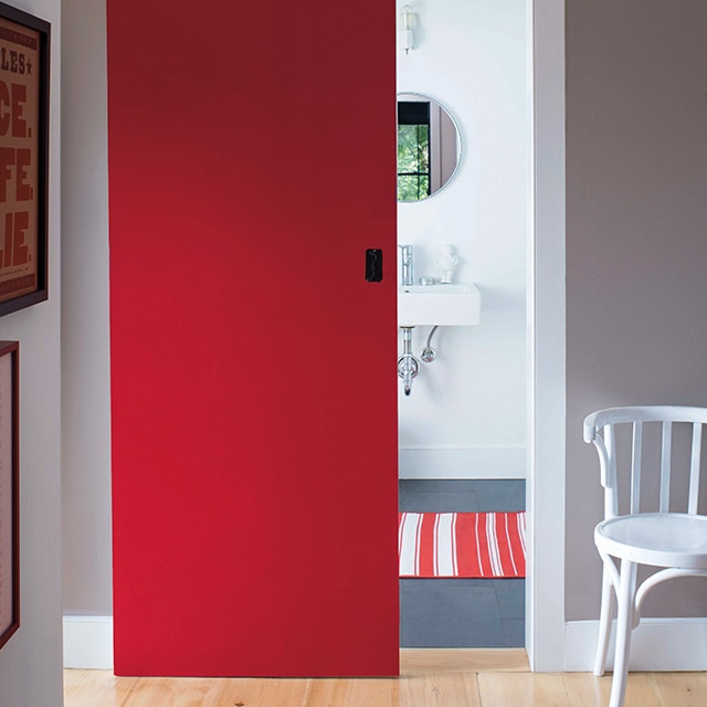 A white spindle chair sits in a gray-walled hallway featuring a red barndoor that leads to a bathroom with a striped rug. A white spindle chair sits in a gray-walled hallway featuring a red barndoor that leads to a bathroom with a striped rug.
