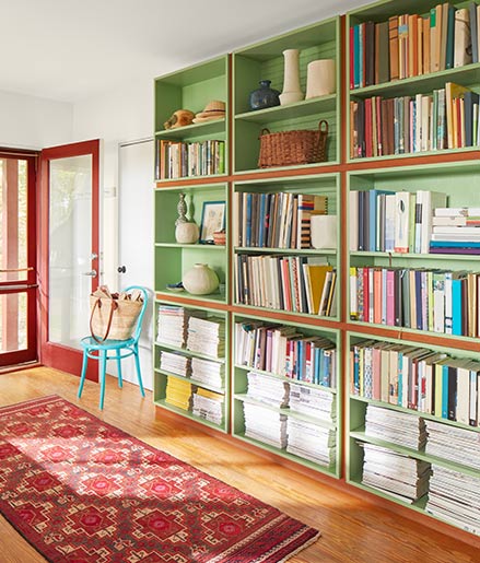 An inviting home entryway with floor-to-ceiling green painted bookshelves, white walls and ceiling, a red-trimmed front door, and a red area rug.
