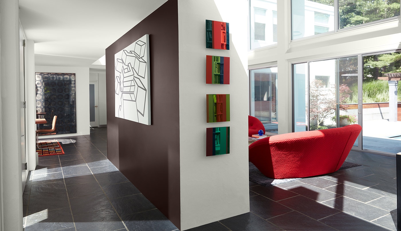 A retro style room with white painted walls and ceiling, a brown accent wall, large windows and a red chair.