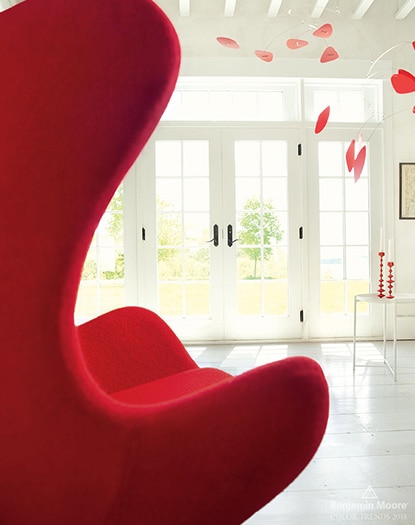 A bright room with white painted walls, beamed ceiling, french doors, red Egg chair and red hanging mobile.