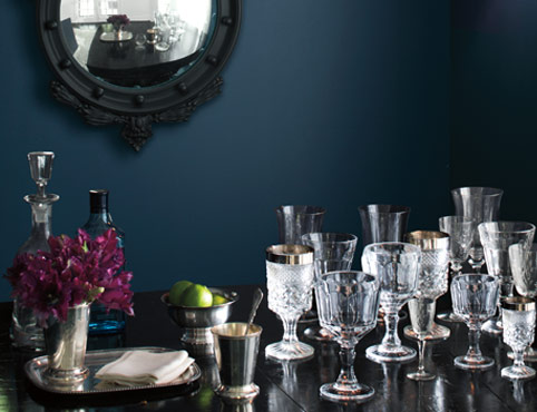 A dark blue-painted dining room wall frames ornate glasses on a table. A dark blue-painted dining room wall frames ornate glasses on a table.