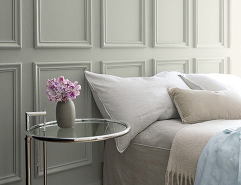 Gray wainscoting behind a bed with gray bedding and a glass side table featuring a small vase of purple flowers. Gray wainscoting behind a bed with gray bedding and a glass side table featuring a small vase of purple flowers.
