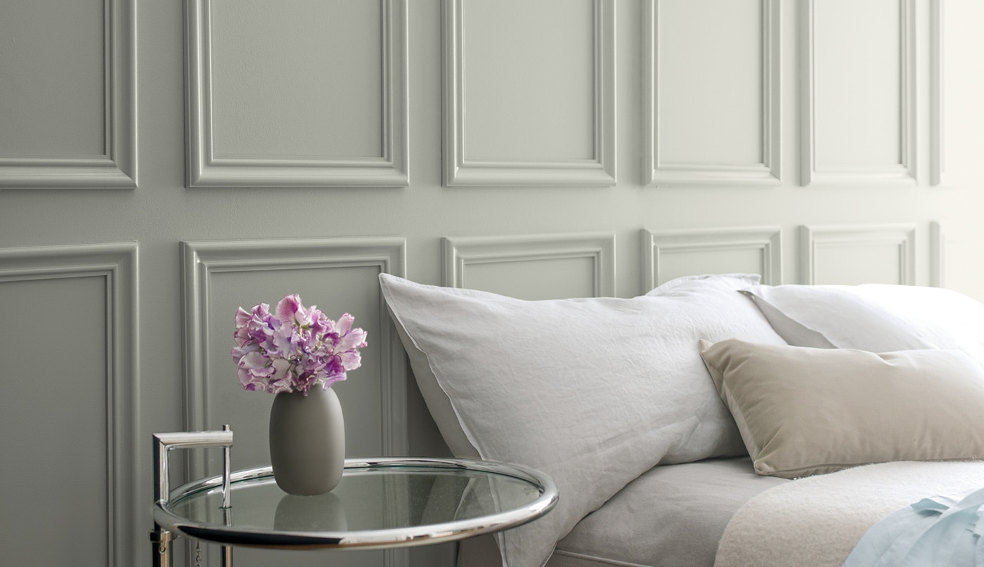 Gray wainscoting behind a bed with gray bedding and a glass side table featuring a small vase of purple flowers.