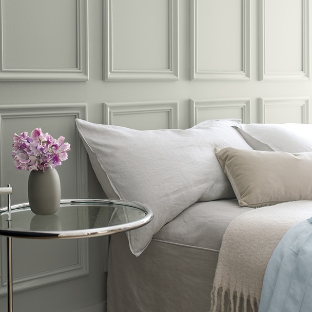 Gray wainscoting behind a bed with gray bedding and a glass side table featuring a small vase of purple flowers.