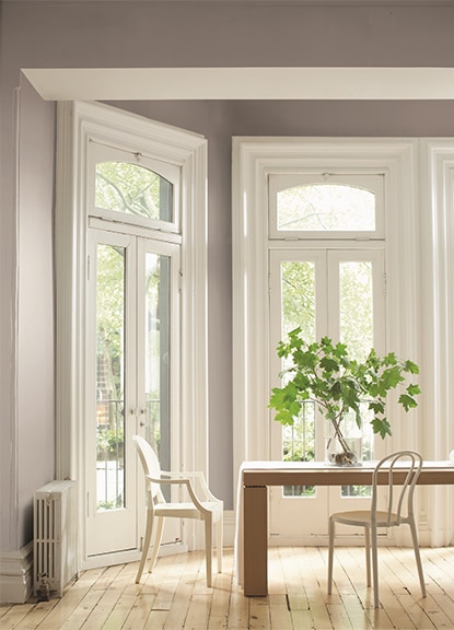 A gray-painted dining room features a wooden plank table, two white chairs, leafy branches in a vase, and French doors. A gray-painted dining room features a wooden plank table, two white chairs, leafy branches in a vase, and French doors.