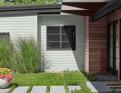 A light, gray-painted modern house with black trim and brown stain, a stone front path, tall grasses, and potted flowers in front. A light, gray-painted modern house with black trim and brown stain, a stone front path, tall grasses, and potted flowers in front.