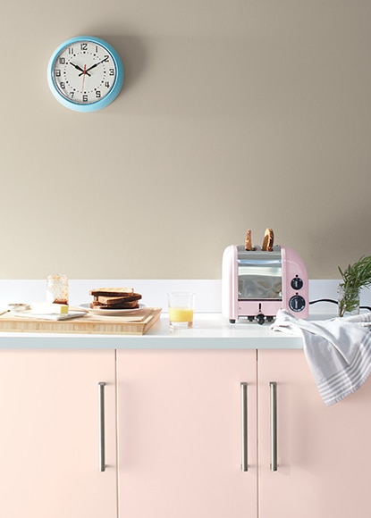 Greige walls with light pink cabinets and white countertops featuring kitchen appliances and a small blue wall clock. Greige walls with light pink cabinets and white countertops featuring kitchen appliances and a small blue wall clock.