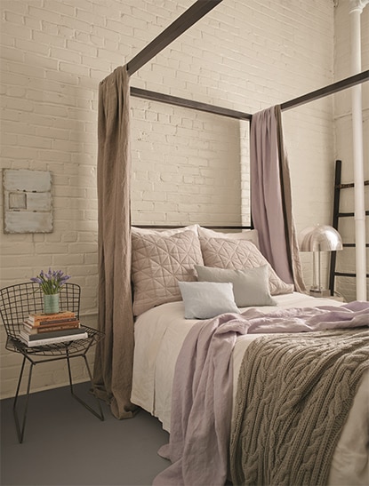 Bedroom with a white washed brick wall behind a tan canopy bed with lilac pillows and throw blanket. Bedroom with a white washed brick wall behind a tan canopy bed with lilac pillows and throw blanket.