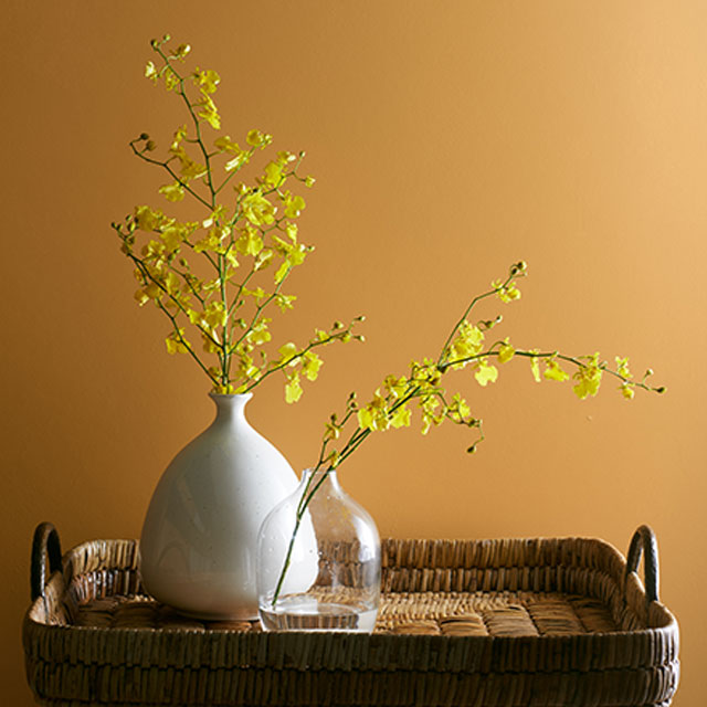 One ceramic vase and one glass vase with yellow flowers on a woven tray in front of an orange-painted wall. One ceramic vase and one glass vase with yellow flowers on a woven tray in front of an orange-painted wall.