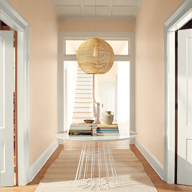 A peach-painted hallway with white doors and trim, rattan ceiling light, modern table, and white rug. A peach-painted hallway with white doors and trim, rattan ceiling light, modern table, and white rug.