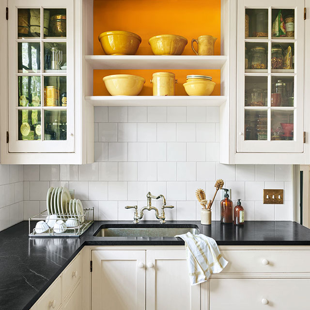 A mostly white-painted kitchen with black counter tops and orange accent behind shelving. A mostly white-painted kitchen with black counter tops and orange accent behind shelving.