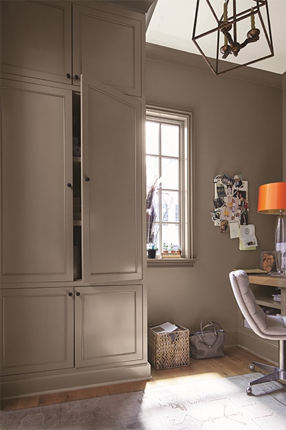 A taupe-painted home office with cupboard, beige chair, wooden desk, note board, white ceiling, and abstract chandelier. A taupe-painted home office with cupboard, beige chair, wooden desk, note board, white ceiling, and abstract chandelier.