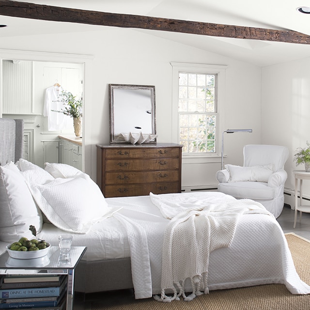 A cozy, white-painted bedroom with white bedding and chairs, a bay window with white cushions, an antique dresser, and a vaulted white ceiling with a dark brown wooden beam, looking into a white bathroom. A cozy, white-painted bedroom with white bedding and chairs, a bay window with white cushions, an antique dresser, and a vaulted white ceiling with a dark brown wooden beam, looking into a white bathroom.
