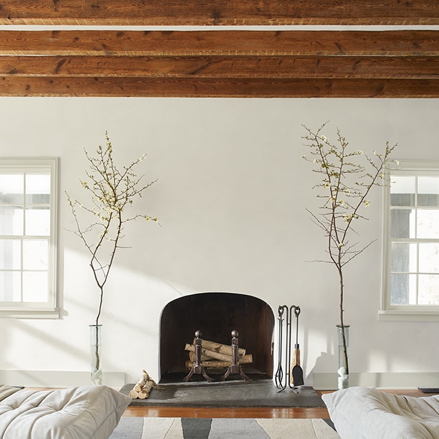 A living room painted in off-white with an arched fireplace flanked by branches in glass vases beneath a ceiling with exposed wood beams. A living room painted in off-white with an arched fireplace flanked by branches in glass vases beneath a ceiling with exposed wood beams.