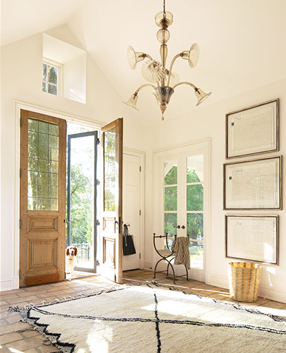 A sunlit, off-white painted entryway with vaulted ceiling, ornate chandelier, a woven beige and black area rug, and a dog looking in from one of two wooden front doors. A sunlit, off-white painted entryway with vaulted ceiling, ornate chandelier, a woven beige and black area rug, and a dog looking in from one of two wooden front doors.