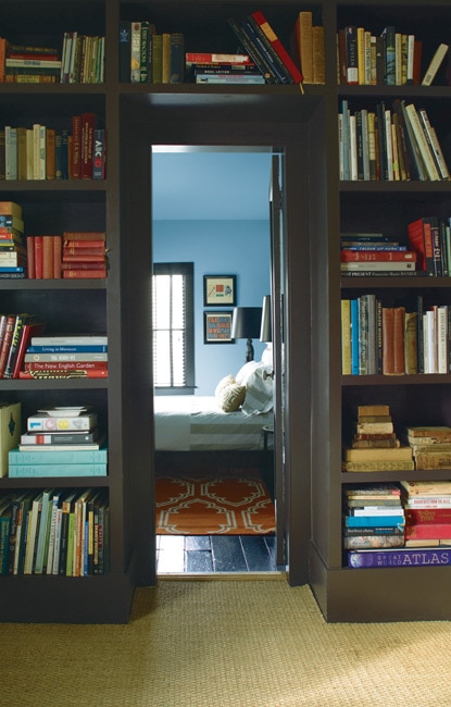 A doorway into a welcoming, blue-hued bedroom lit by natural light is framed by dark wood shelving featuring a large collection of books. A doorway into a welcoming, blue-hued bedroom lit by natural light is framed by dark wood shelving featuring a large collection of books.