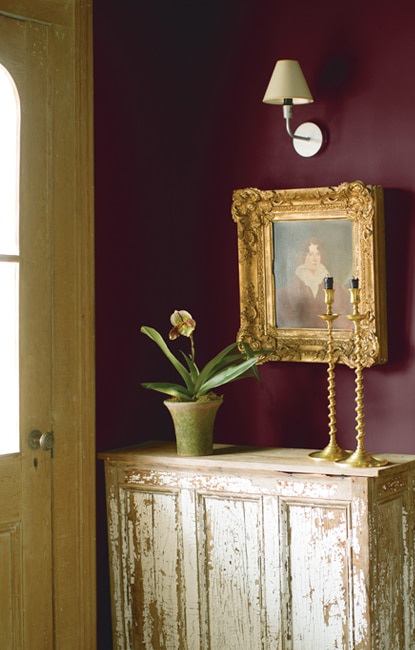This light-flooded entryway features two tapered candles and a potted flower on top of a weathered antique table set against a deep, plum-colored wall. This light-flooded entryway features two tapered candles and a potted flower on top of a weathered antique table set against a deep, plum-colored wall.