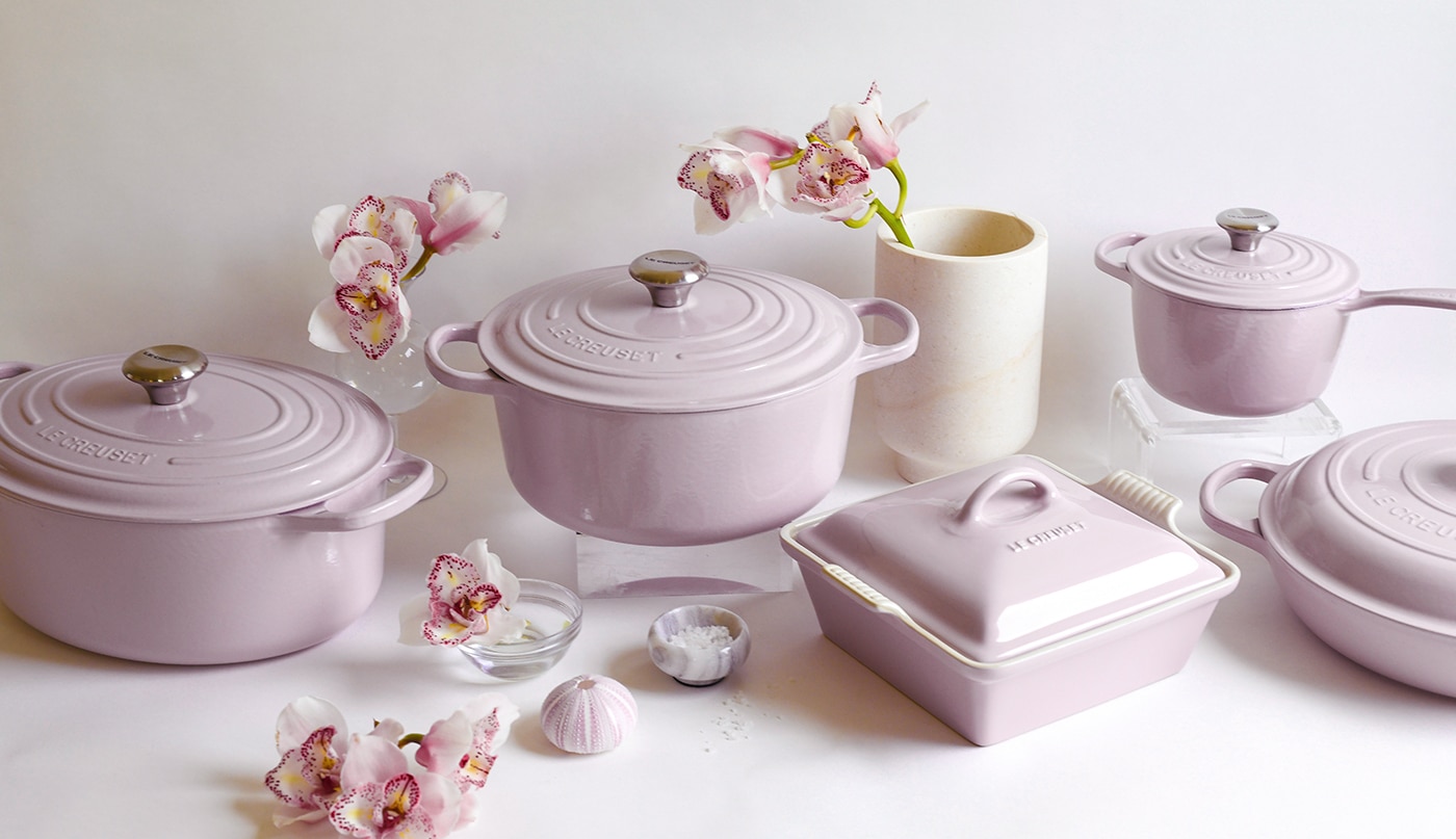 An array of Le Creuset cookware in Shallot, the newest Le Creuset color, a lovely lavender-gray, alongside several fresh flowers set off from off-white wall.
