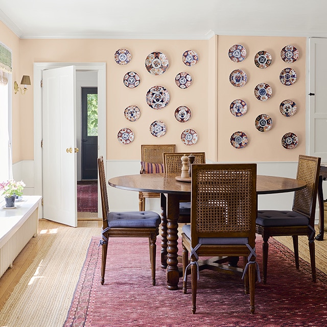 Dining room with pale pink on the upper portion of the walls and white on the lower portion; a wooden bench is set beneath large, bright windows that illuminate a wooden dining table set on a red rug and light wood flooring. Dining room with pale pink on the upper portion of the walls and white on the lower portion; a wooden bench is set beneath large, bright windows that illuminate a wooden dining table set on a red rug and light wood flooring.