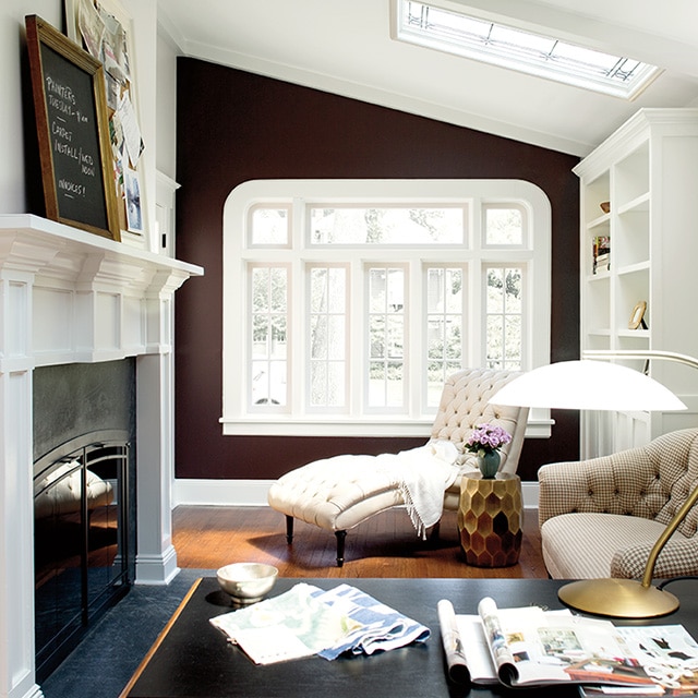 A study with white walls and a chocolate brown accent wall set with a large, white-framed window, a fireplace, a desk and several other pieces of furniture spread throughout the room. A study with white walls and a chocolate brown accent wall set with a large, white-framed window, a fireplace, a desk and several other pieces of furniture spread throughout the room.
