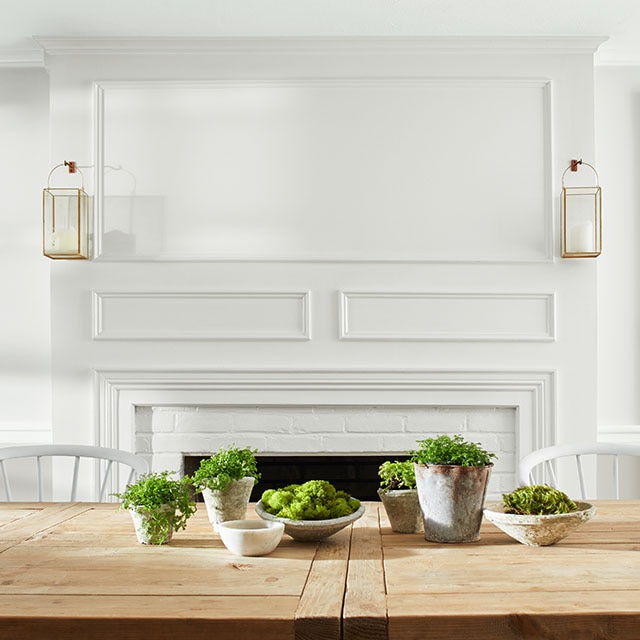 An elegant white dining room with white-painted brick fireplace with lanterns above mantel, and wooden table with several potted succulents on top of it. An elegant white dining room with white-painted brick fireplace with lanterns above mantel, and wooden table with several potted succulents on top of it.