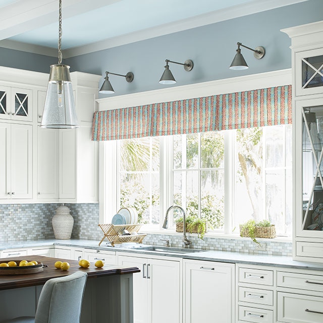 A welcoming kitchen with light-blue painted walls and ceiling painted in an icy blue framing white beams, three large windows over a sink, a kitchen island, and white-painted cabinetry. A welcoming kitchen with light-blue painted walls and ceiling painted in an icy blue framing white beams, three large windows over a sink, a kitchen island, and white-painted cabinetry.